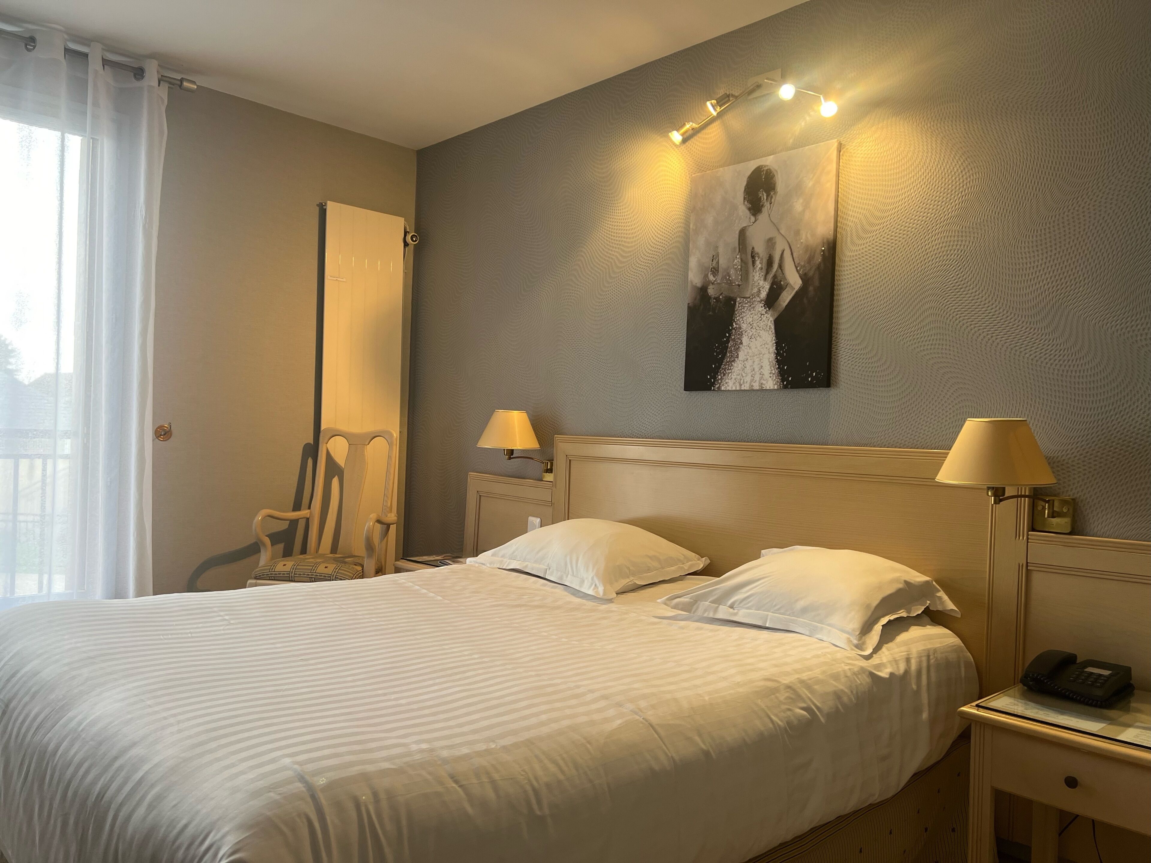 Ein Hotelzimmer mit einem großen Bett, zwei Nachttischlampen, einem Sessel, einem Bild an der Wand und einem Telefon auf dem Nachttisch.