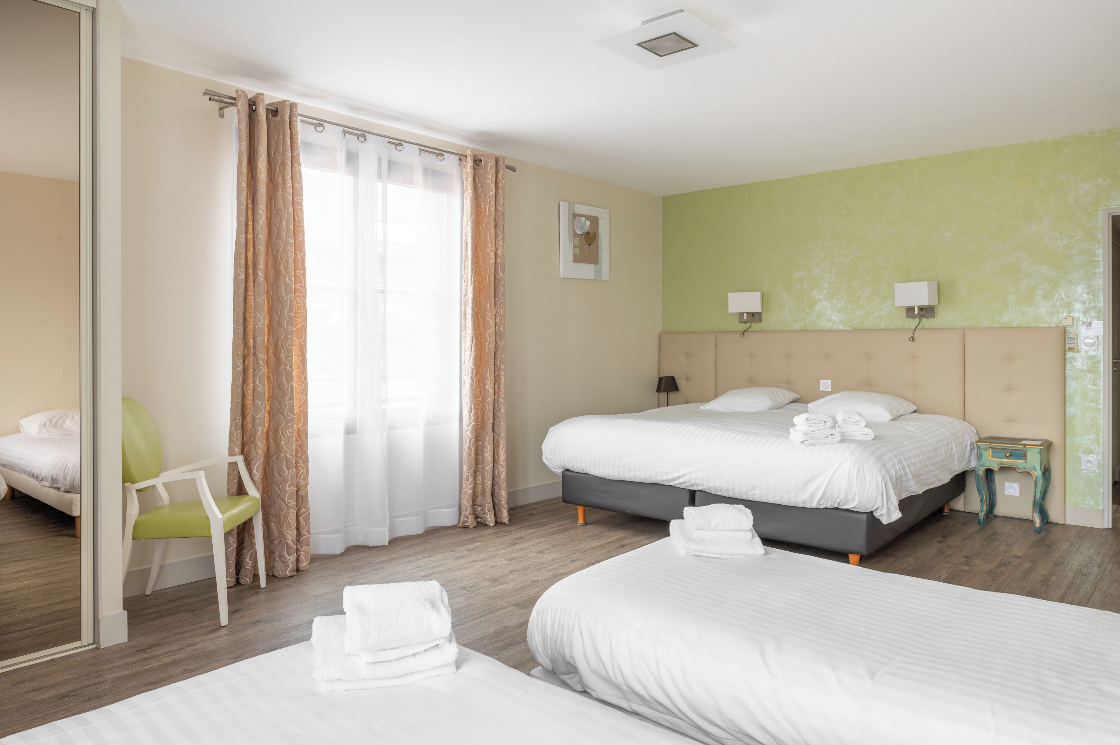 Une chambre d’hôtel avec deux lits, un fauteuil vert, une petite table et un miroir.