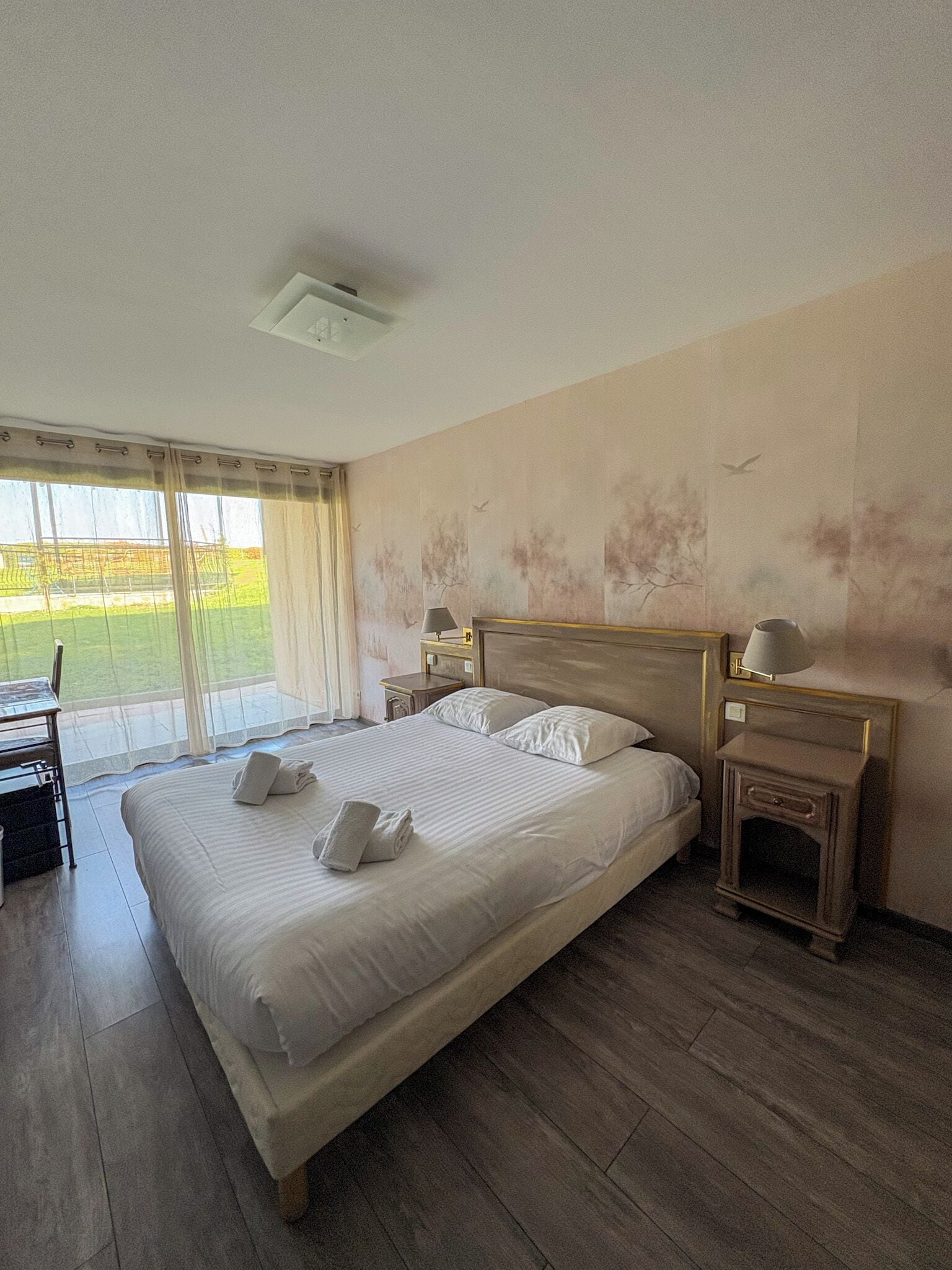 Ein Hotelzimmer mit einem großen Bett, zwei Nachttischen, einem Sessel und Blick ins Freie.