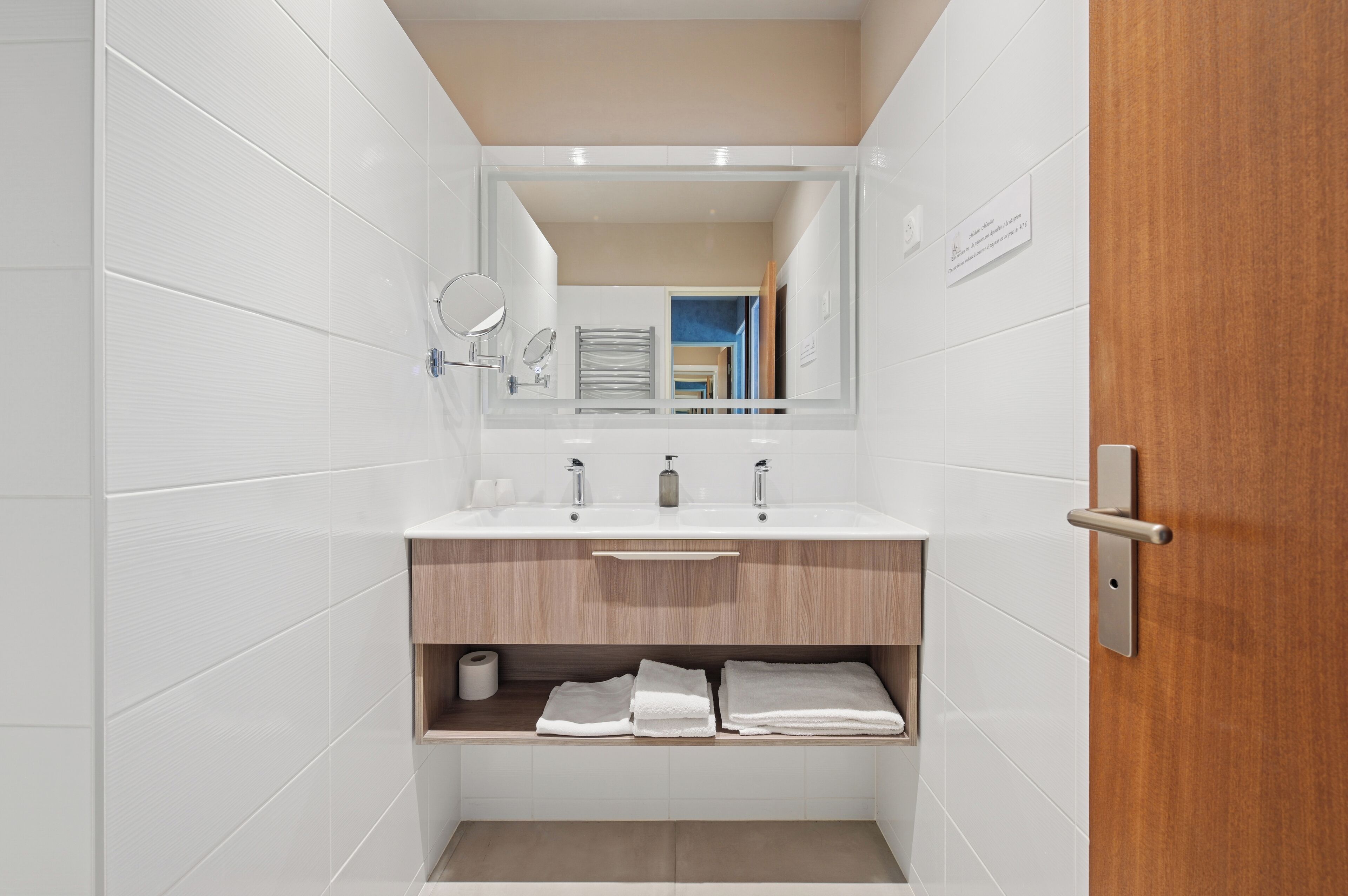 A modern bathroom with white tiles, a wooden vanity, a mirror, and a towel rack.