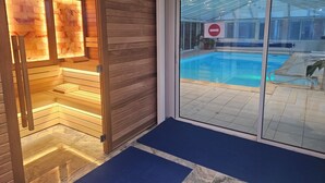 Un área de piscina interior moderna con puerta de cristal que da a una piscina, bancos de madera y un tapete azul en la entrada.