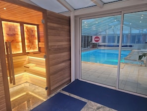 Een moderne binnenzwembadruimte met een glazen deur naar het zwembad, houten bankjes en een blauwe mat bij de ingang.