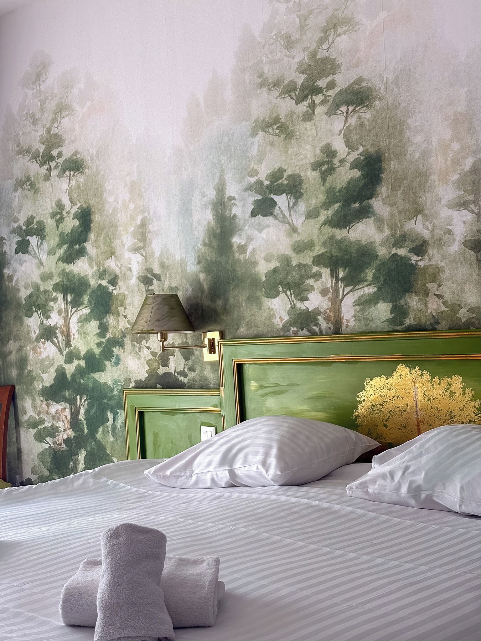 A neatly made bed with white linens and a folded towel, a green headboard, and a wall with a painted tree mural.