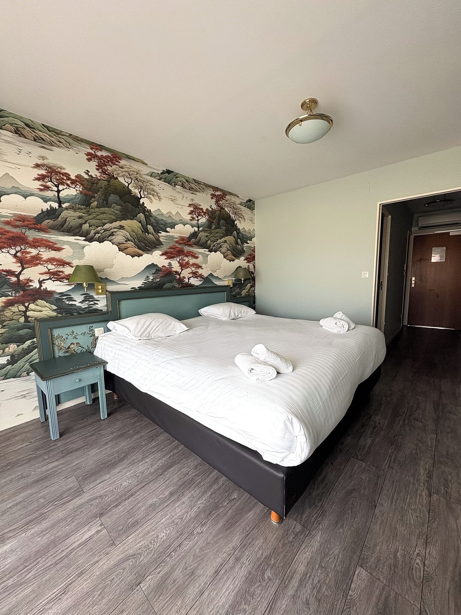 A bedroom with a bed, a nightstand, a wall mural, and a door.