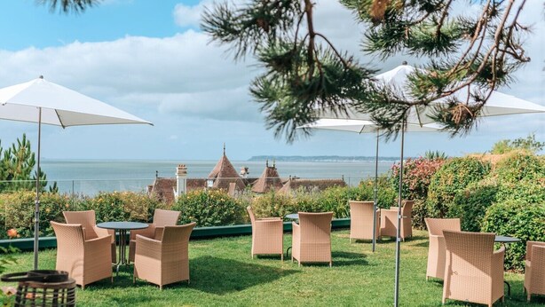 An outdoor seating area with wicker chairs and tables, overlooking a body of water and buildings in the background. - Hotel Restaurant Le Bellevue (Villerville)