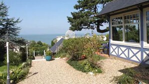 A house with a gravel pathway leading to a blue door, surrounded by greenery and a view of the sea. - Hotel Restaurant Le Bellevue (Villerville)