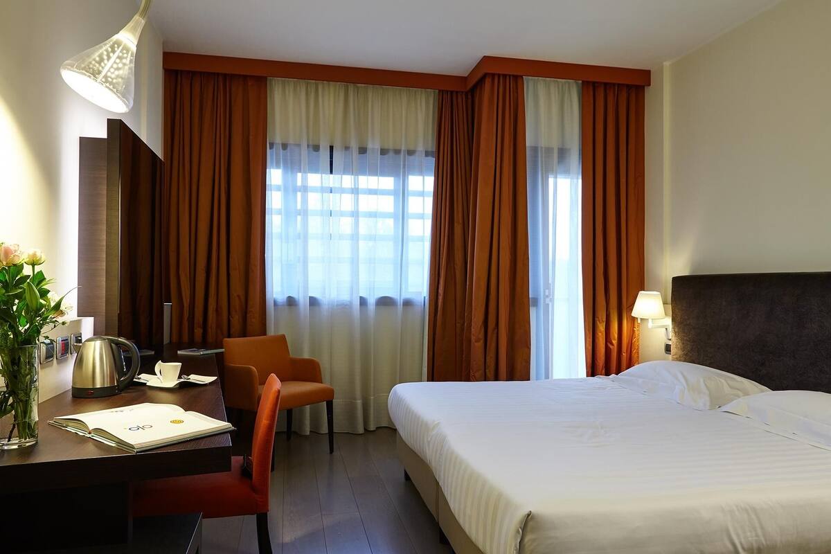 Habitación de hotel con cama, escritorio, silla, lámpara y ventana con cortinas.