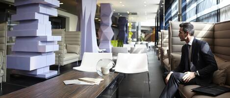 Un moderno vestíbulo de hotel con un hombre sentado en un sofá, una mesa de madera con una cúpula de cristal y mobiliario contemporáneo.