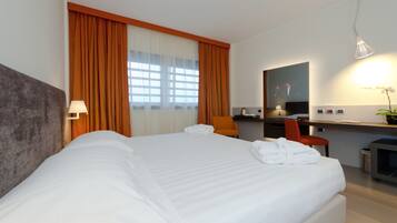 Une chambre d’hôtel avec un grand lit, un bureau avec une chaise, une petite table, une plante en pot et un miroir au mur.