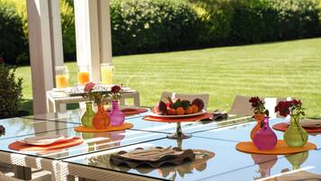 Un espace de restauration en plein air avec une table en verre, des chaises en osier et une variété d’éléments de décoration de table, notamment des assiettes, des couverts et des vases avec des fleurs.
