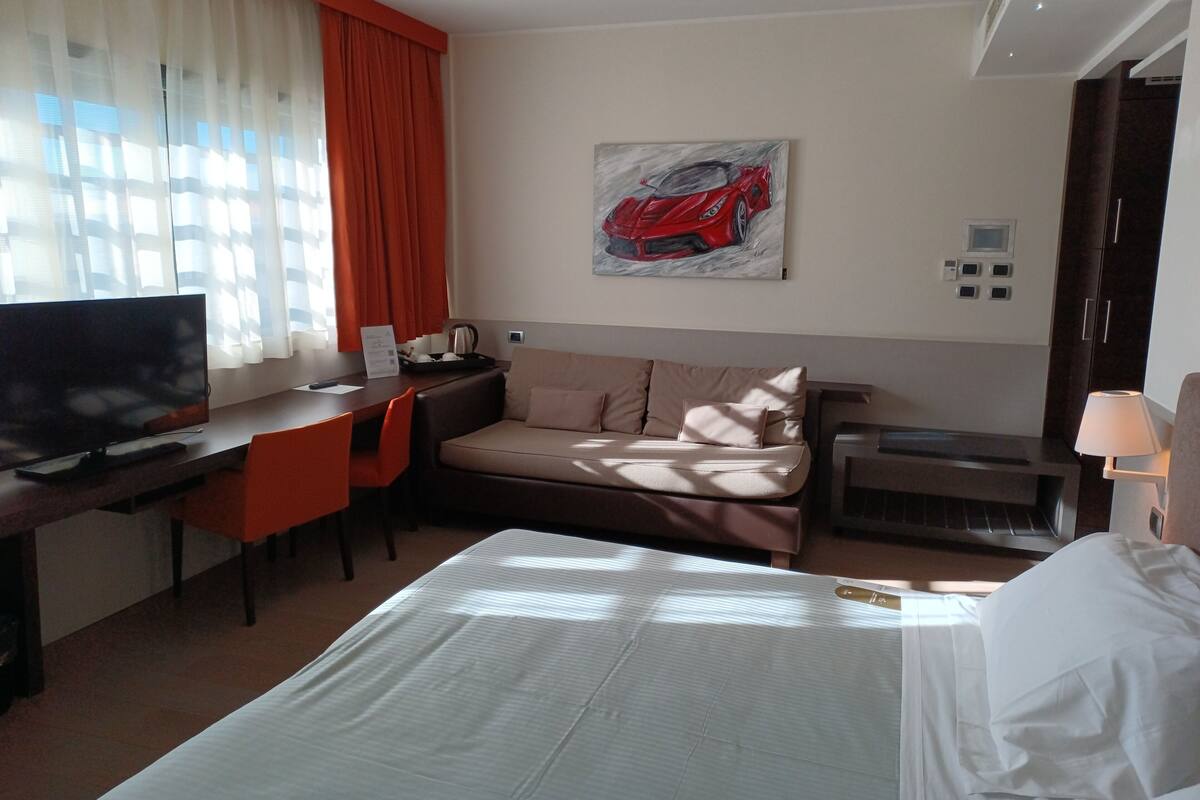 Una habitación de hotel con una cama, un escritorio con televisor, un sofá y un cuadro de un coche en la pared.