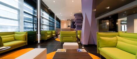 A modern lounge area with green sofas and a central table, featuring magazines and a flat-screen TV.