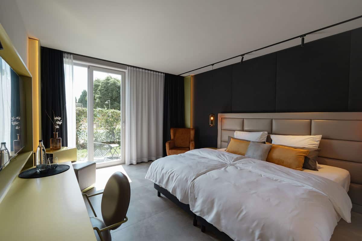 Una habitación de hotel moderna con una cama grande, un escritorio y una silla. Hay una ventana con cortinas y vistas a la vegetación.