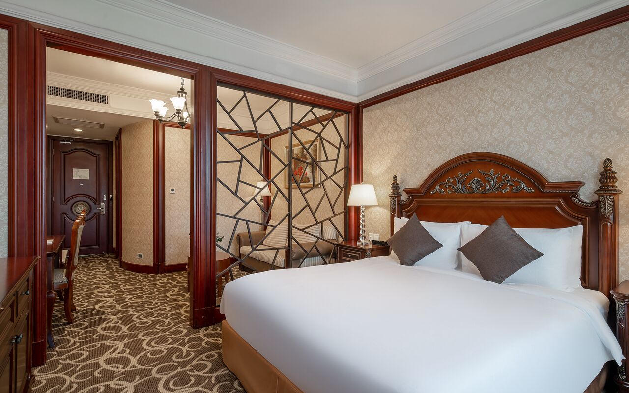 A hotel room with a large bed, a wooden headboard, a nightstand, a lamp, and a patterned carpet.