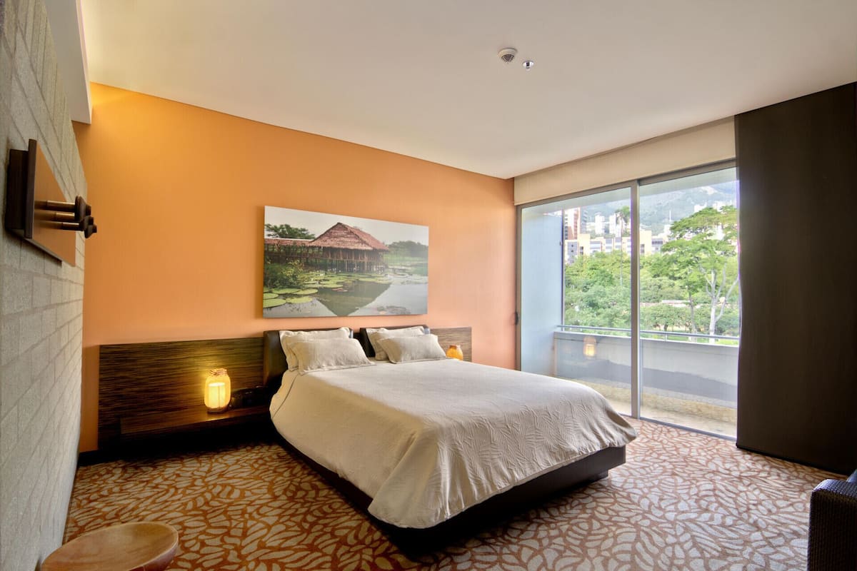 A hotel room with a bed, a large window, a painting on the wall, and a nightstand with a lamp.