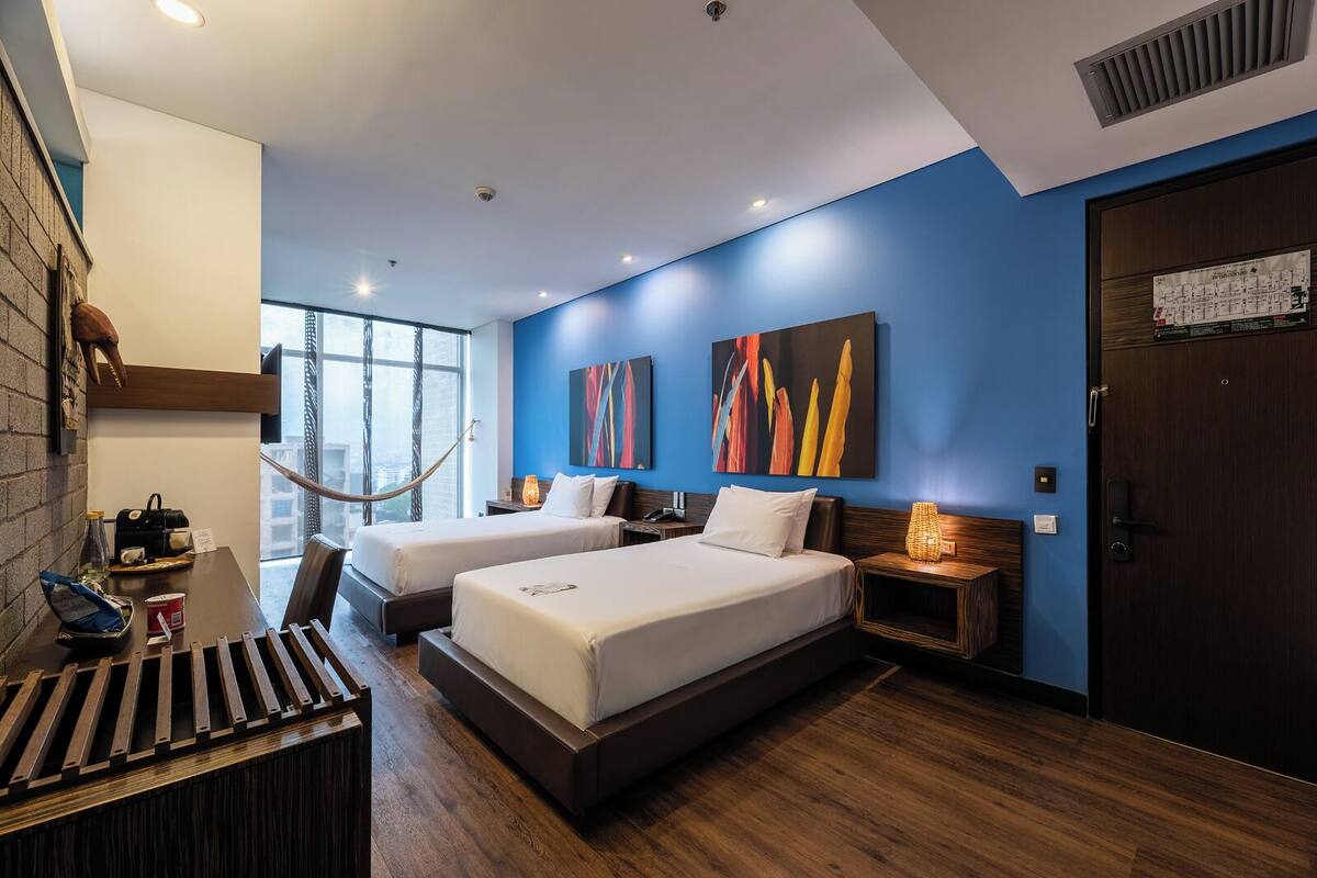 A hotel room with two beds, a hammock, a wooden desk, and a large window.