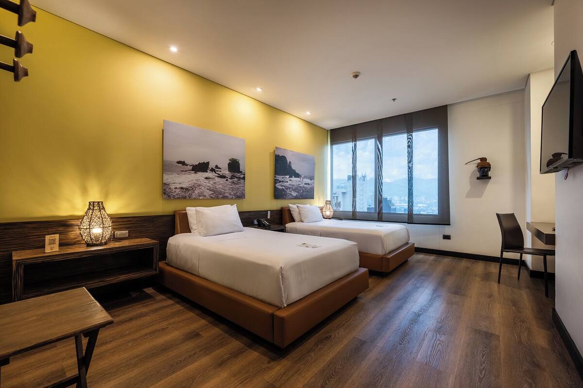 A hotel room with two beds, a desk, a chair, and a large window with a city view.