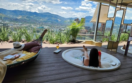 A person relaxing in a hot tub with a view of mountains and a cityscape.