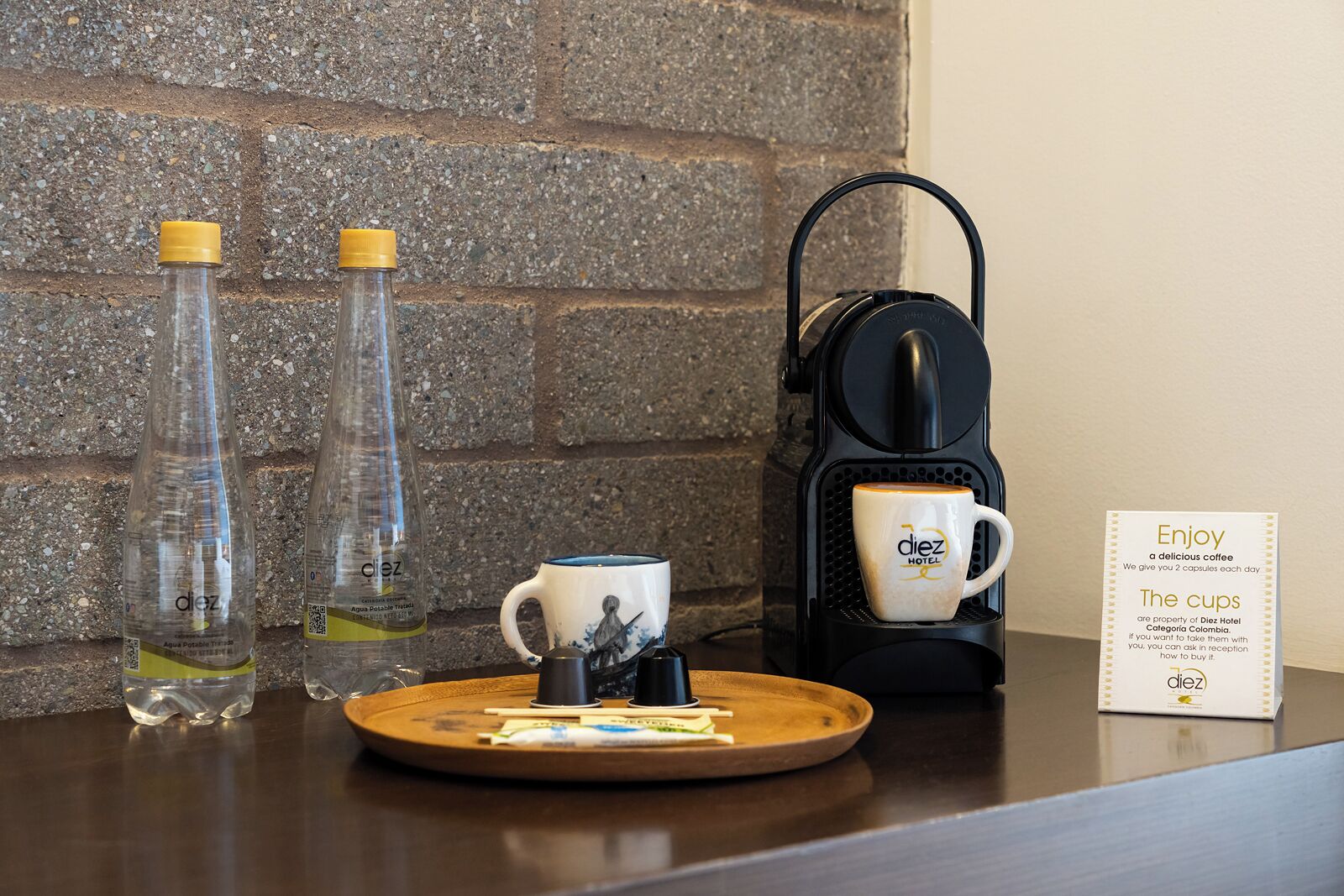 A coffee maker, two water bottles, a mug, and a sign on a counter with a brick wall background.