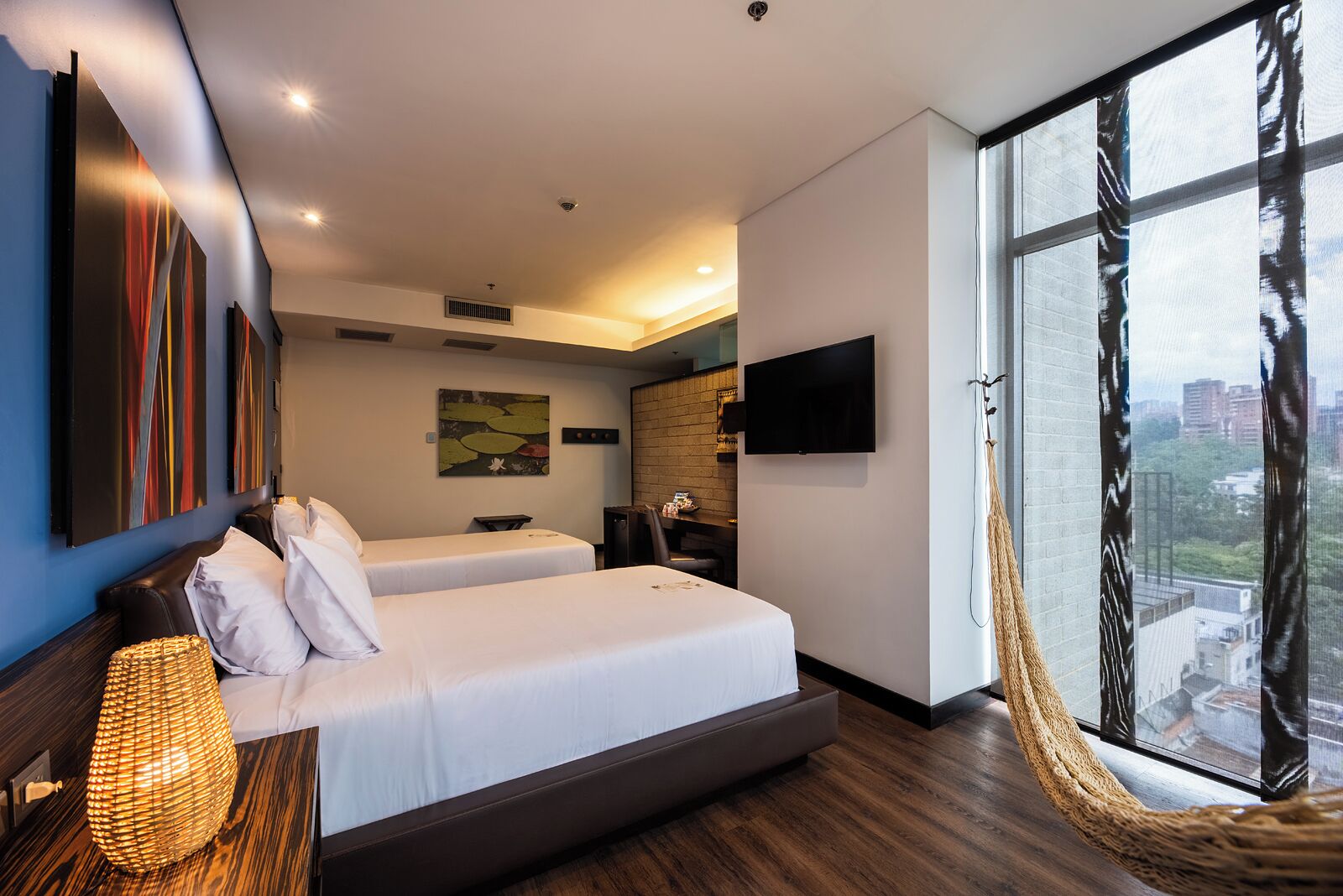 A modern hotel room with two beds, a large window with a view, a flat-screen TV, and artwork on the walls.