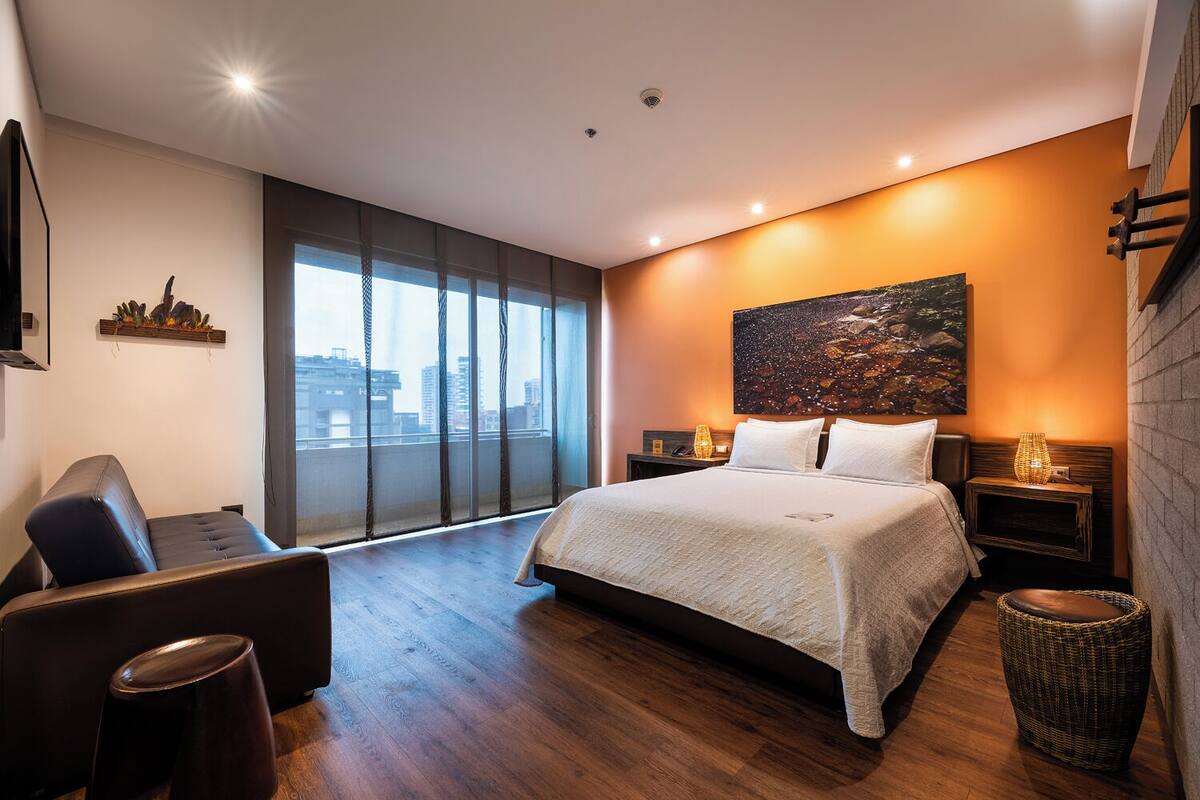 A modern hotel room with a large bed, a seating area, a city view, and a large artwork on the wall.