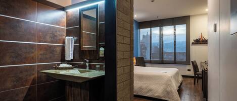 Un baño moderno con un espejo grande, un lavamanos y tocador. Se ve una cama con vista a la ciudad al fondo.