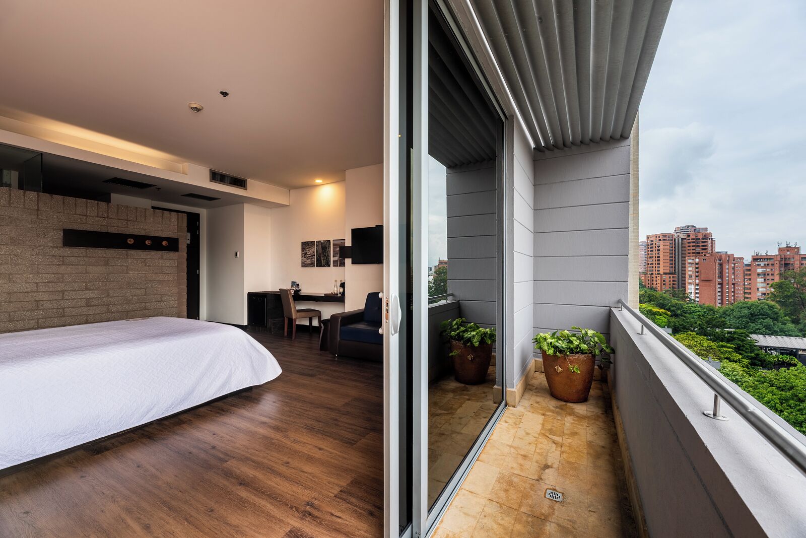 Un dormitorio moderno con una cama grande, un escritorio y una silla, un balcón con plantas en macetas y vistas a la ciudad.