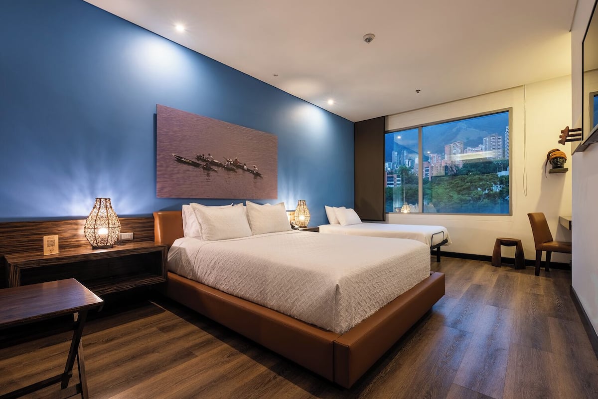 A hotel room with a large bed, two bedside tables, a nightstand, a chair, a large window with a city view, and a wall-mounted artwork.