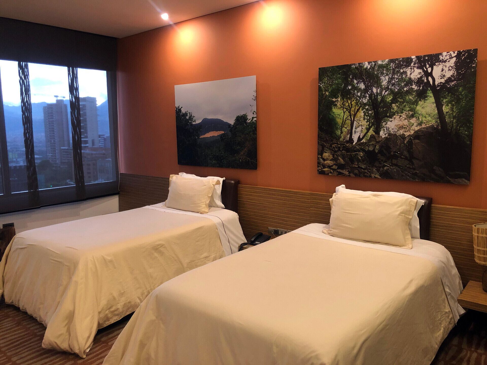 Une chambre d’hôtel avec deux lits, une grande fenêtre donnant sur la ville et deux tableaux accrochés au mur.