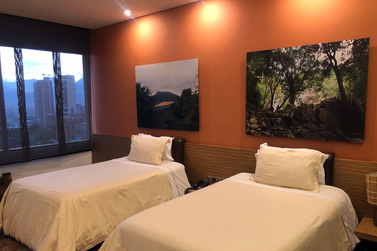 A hotel room with two beds, a large window with a city view, and two paintings on the wall.