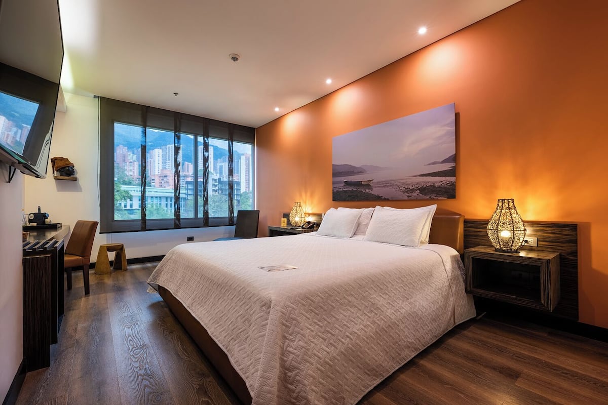 A modern hotel room with a large bed, a desk, a chair, a TV, and a window with a city view.