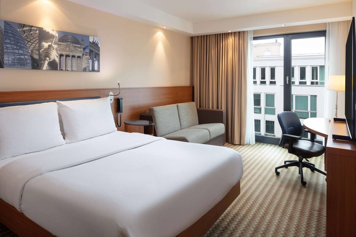 Une chambre d’hôtel avec un grand lit, un canapé, un bureau avec une chaise et une vue sur les immeubles par la fenêtre.