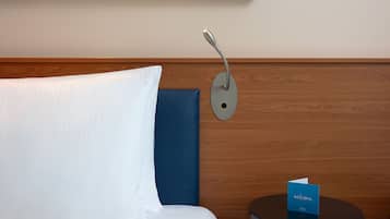 Habitación de hotel con cama, almohada azul, luz nocturna en la pared y una tarjeta de bienvenida sobre una mesita.
