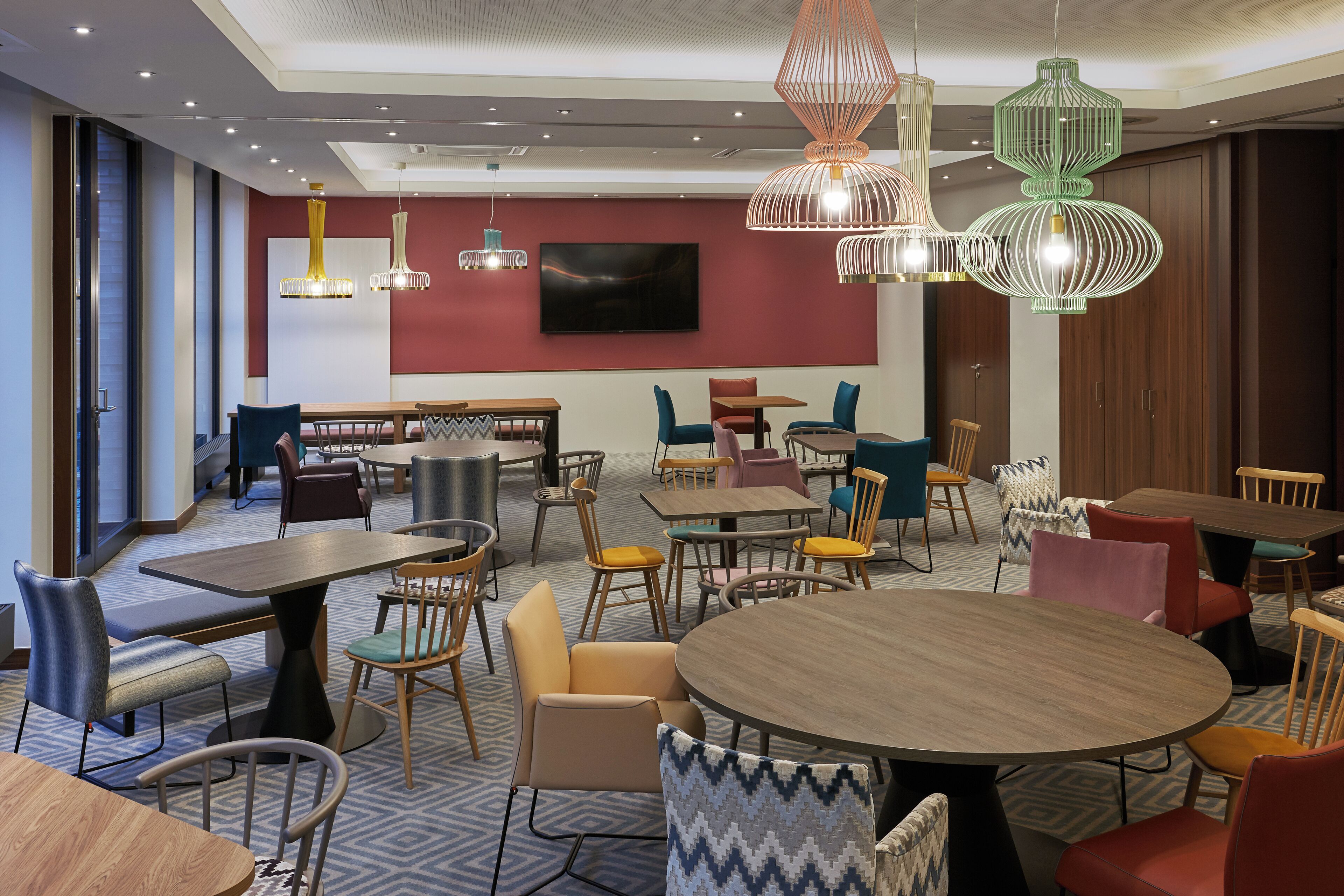 Un espace repas moderne doté de plusieurs tables et chaises, d’un téléviseur à écran plat fixé au mur et de luminaires décoratifs suspendus.