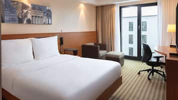 A hotel room with a large bed, a desk with a chair, a small table, a telephone, and a window with curtains.