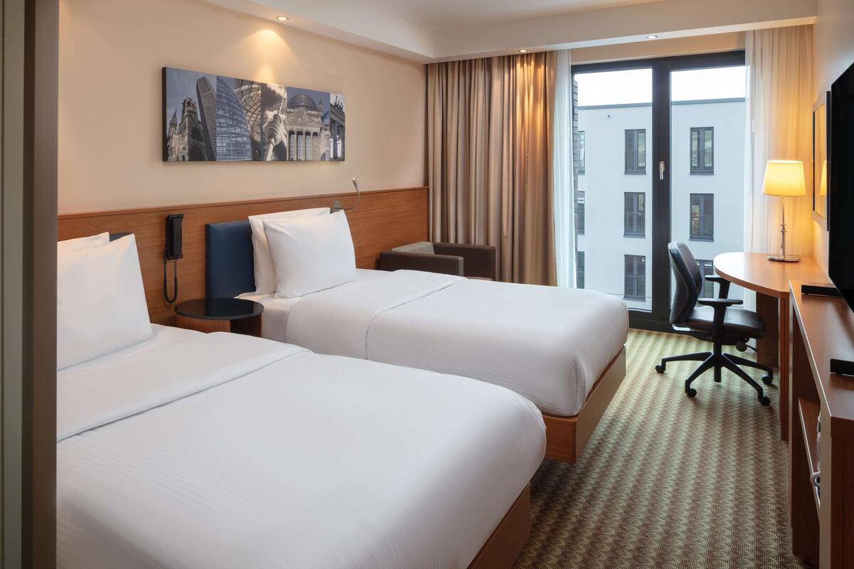 Une chambre d’hôtel avec deux lits, un bureau avec une chaise, un téléphone et un tableau au mur.