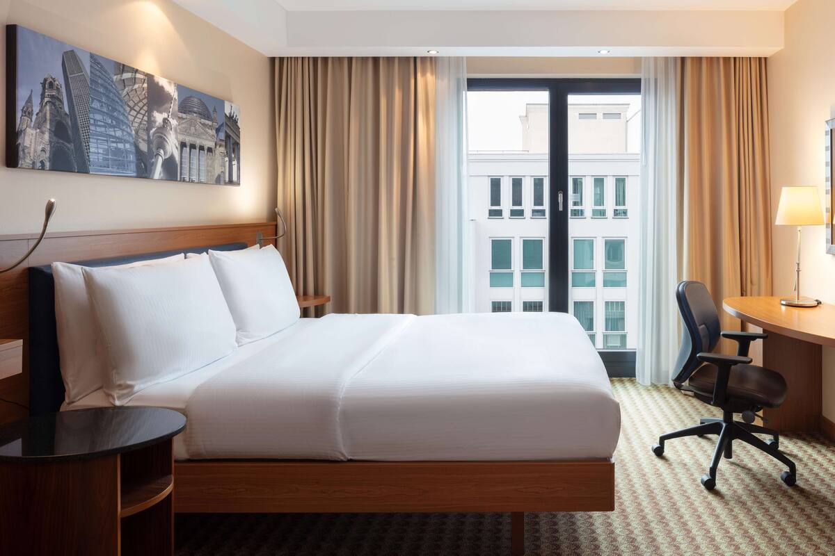 Une chambre d’hôtel avec un grand lit, un bureau, une chaise, une table de chevet et une fenêtre avec des rideaux.