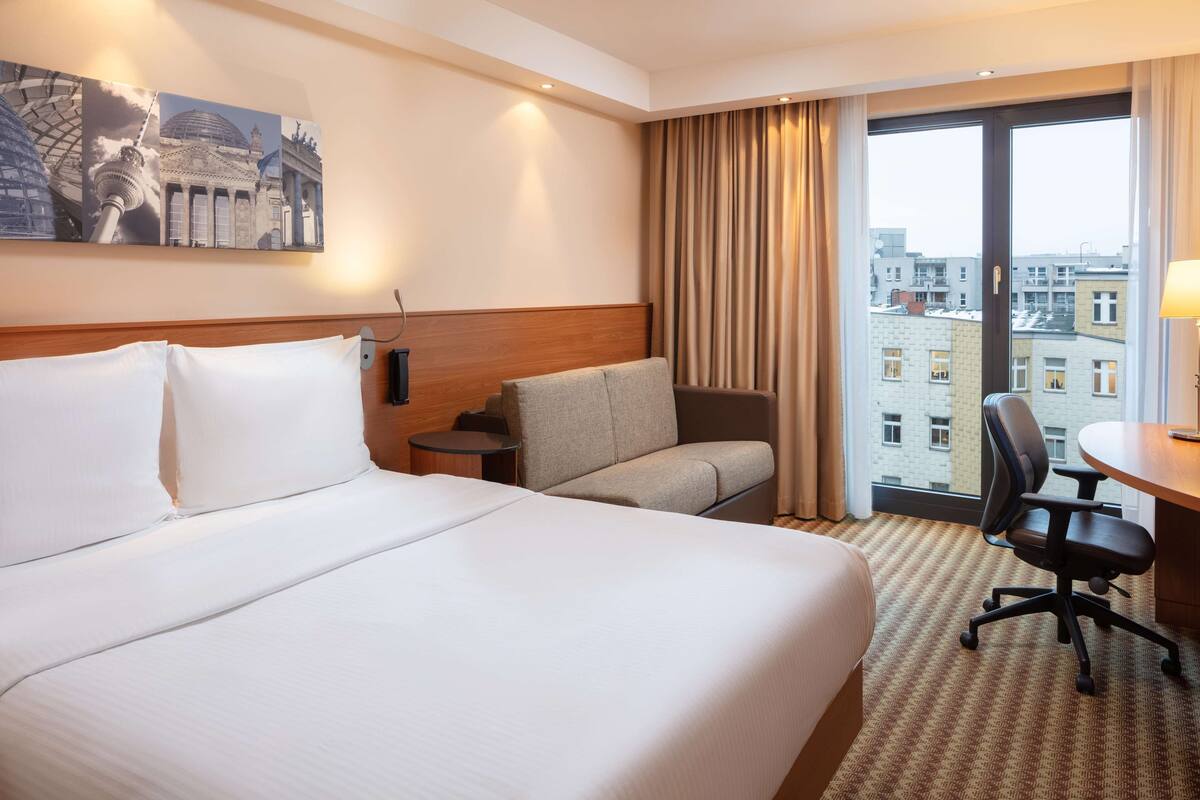 Une chambre d’hôtel avec un grand lit, un canapé, un bureau et une chaise. Il y a un tableau au mur et, par la fenêtre, on peut voir des bâtiments.