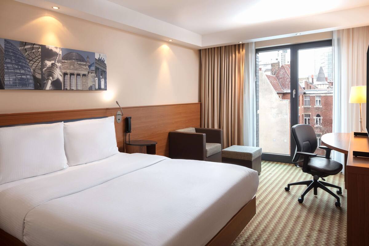 Une chambre d’hôtel avec un grand lit, un bureau, une chaise et une fenêtre donnant sur des immeubles.