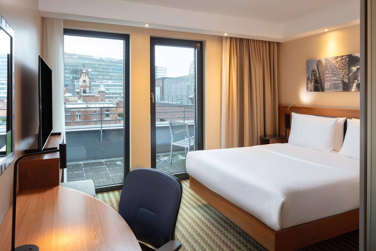 Une chambre d’hôtel avec un grand lit, un bureau, une chaise, une télévision et un balcon donnant sur les immeubles.