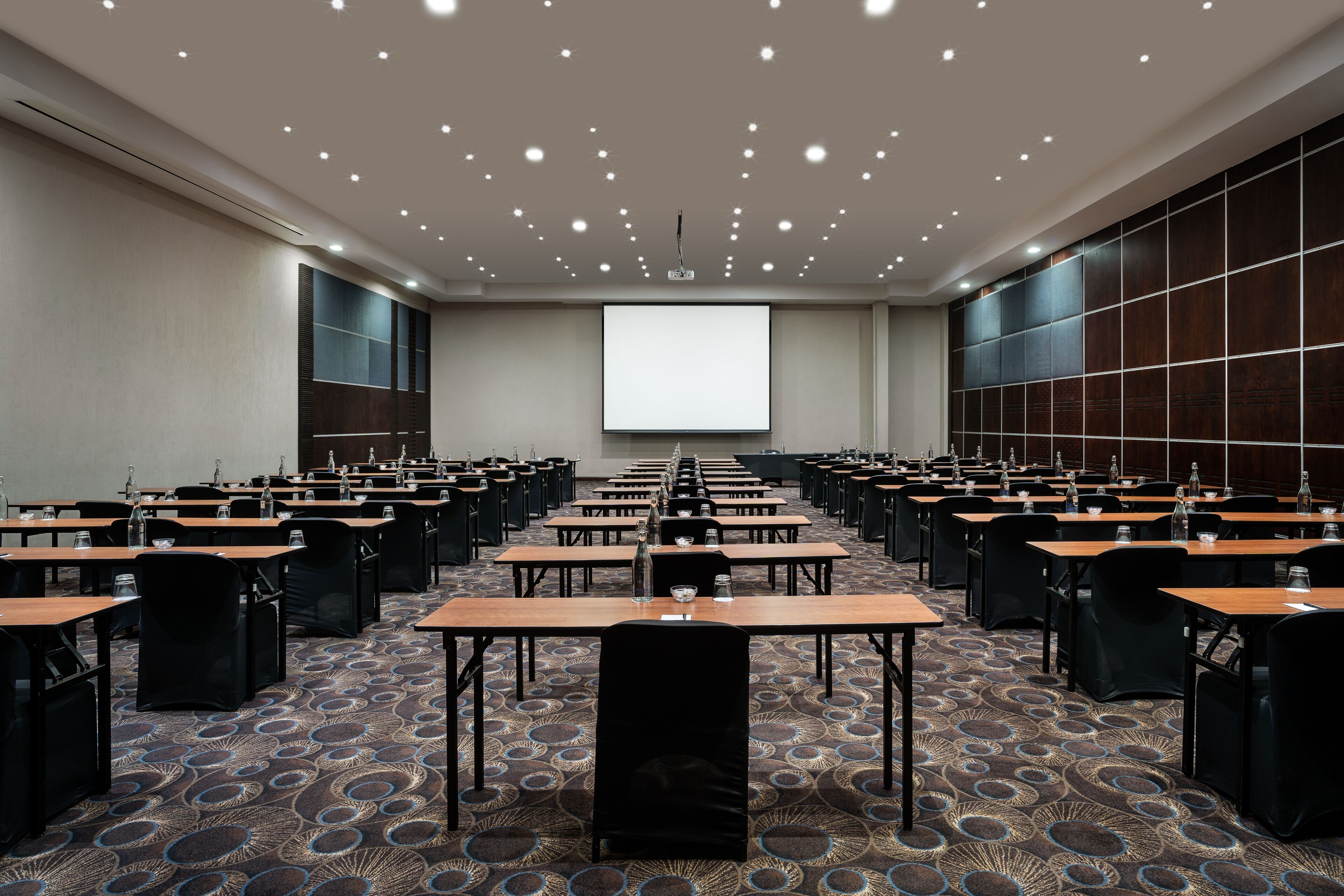 A conference room with rows of tables and chairs, a large screen at the front, and a patterned carpet.