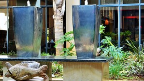 Un espacio exterior moderno con dos grandes fuentes de agua, una palmera y un edificio con amplias ventanas al fondo.