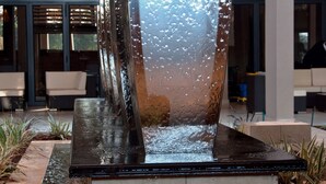 Una fuente de agua interior moderna con superficie reflectante, rodeada de plantas en macetas y un área de descanso con una escalera.