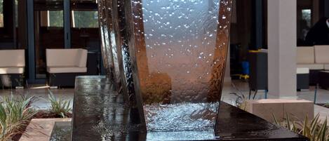 Una fuente de agua interior moderna con superficie reflectante, rodeada de plantas en macetas y un área de descanso con una escalera.