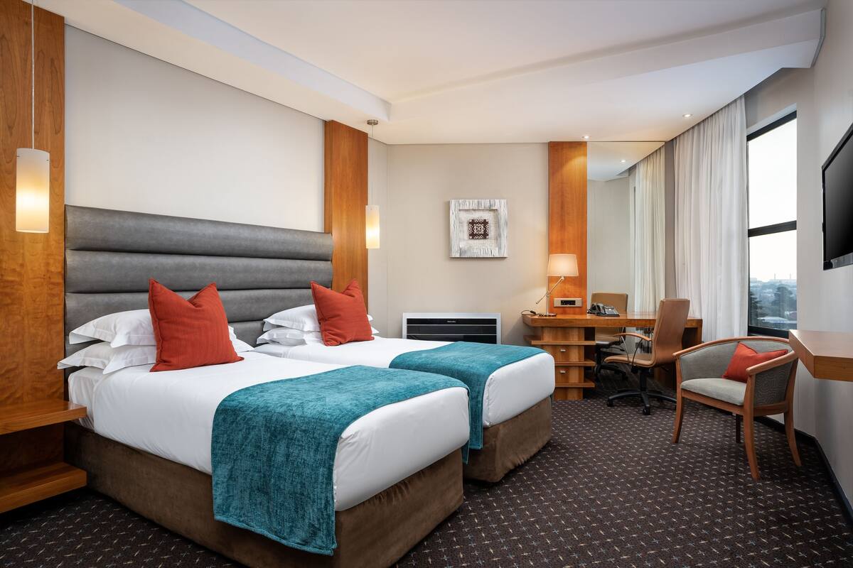 A hotel room with two beds, a desk, a chair, a TV, and a large window.