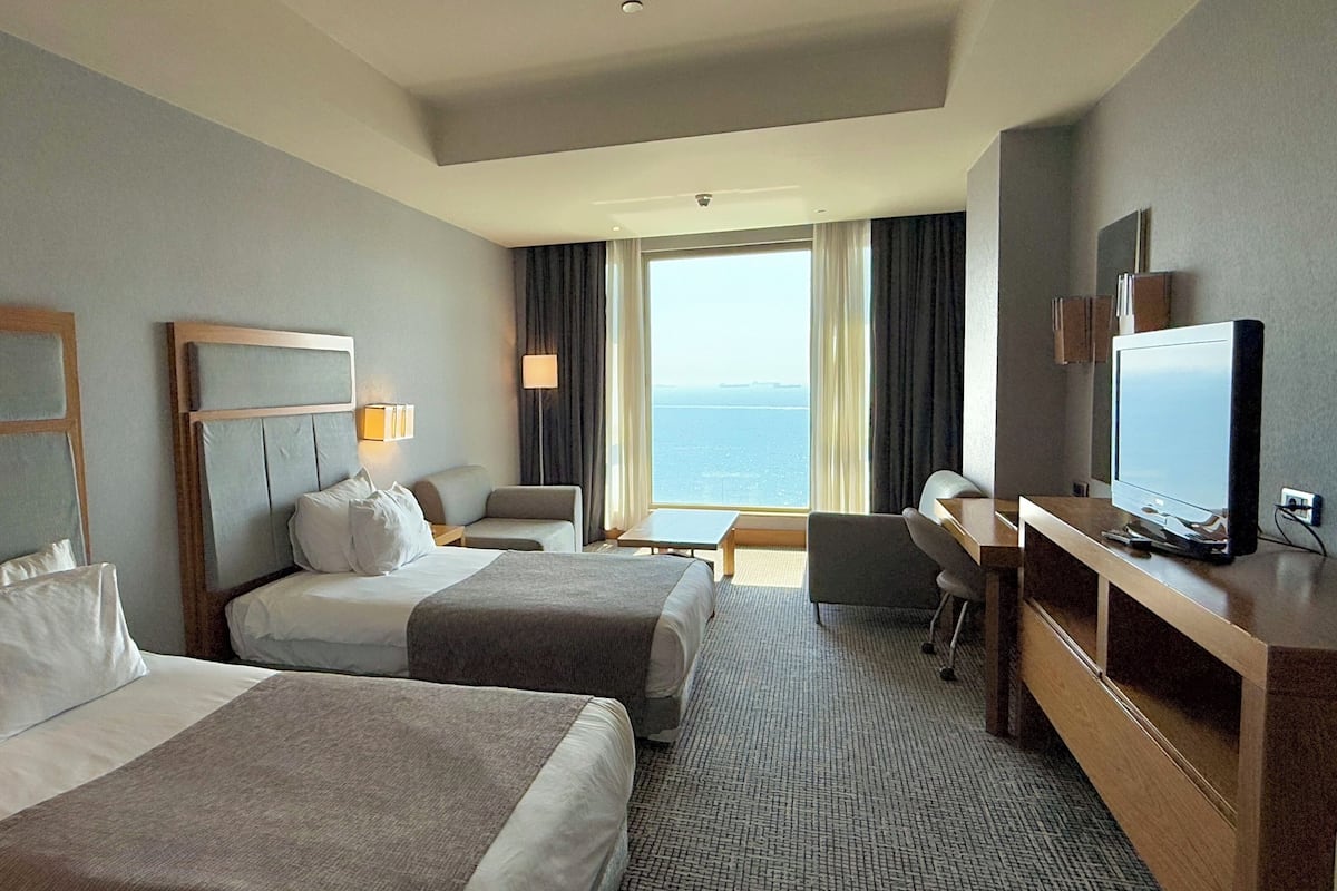 Ein Hotelzimmer mit zwei Betten, einem Schreibtisch, einem Sessel, einem Fernseher und einem großen Fenster mit Blick auf das Meer.