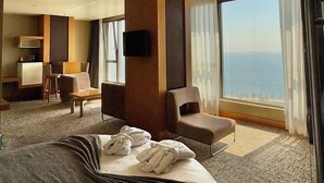 Habitación de hotel con una cama grande, un escritorio con silla, un sofá, un televisor montado en la pared y un ventanal con vistas al océano.