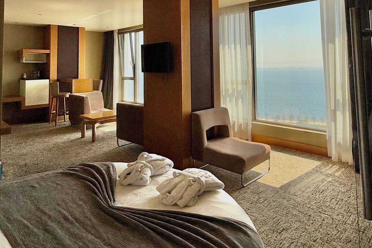 Ein Hotelzimmer mit einem großen Bett, einem Schreibtisch mit Stuhl, einer Sitzecke und einem Fernseher an der Wand. Ein großes Fenster bietet Blick aufs Meer.