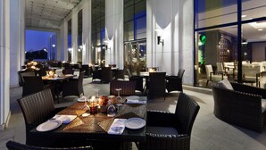 Un área de comedor al aire libre con mesas dispuestas para dos, rodeada de sillas de mimbre y con velas.
