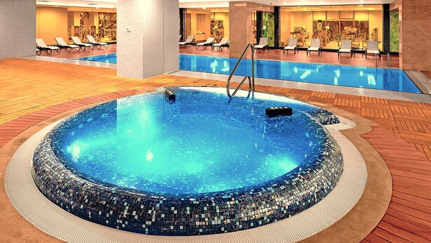 Una amplia piscina cubierta con borde de mosaicos, rodeada de sillas de descanso y un área de gimnasio al fondo.